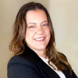 Retrato de Monisa Reis, CEO da Sacratu, sorrindo, de blazer preto sobre fundo bege.