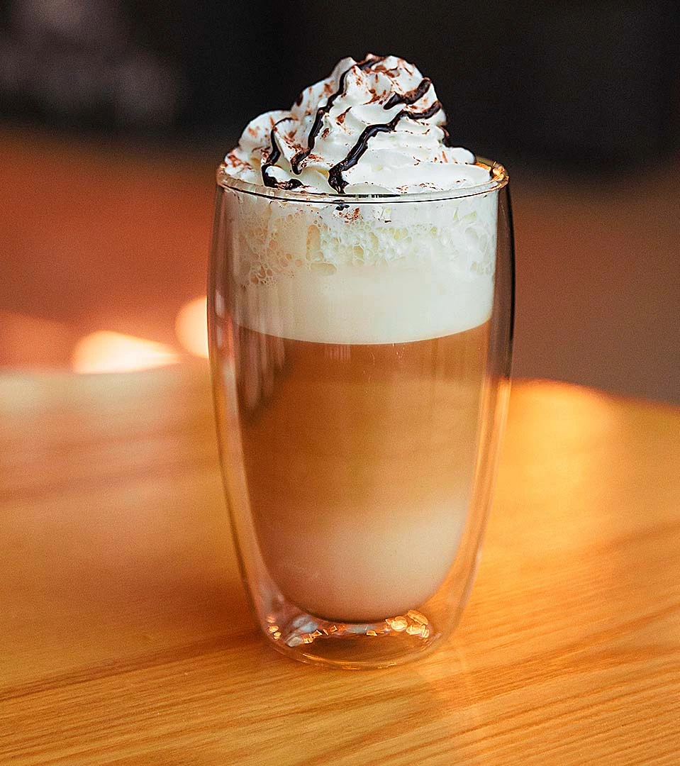 Copo de vidro de parede dupla com café, leite vaporizado e chantilly com calda de chocolate, sobre um balcão de madeira.