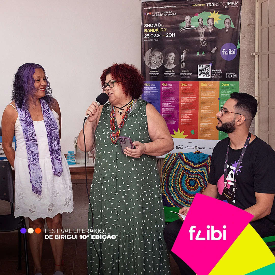 Abertura do Flibi na biblioteca: uma mulher fala ao microfone entre duas pessoas sentadas, diante do cartaz com a programação completa dos cinco dias.