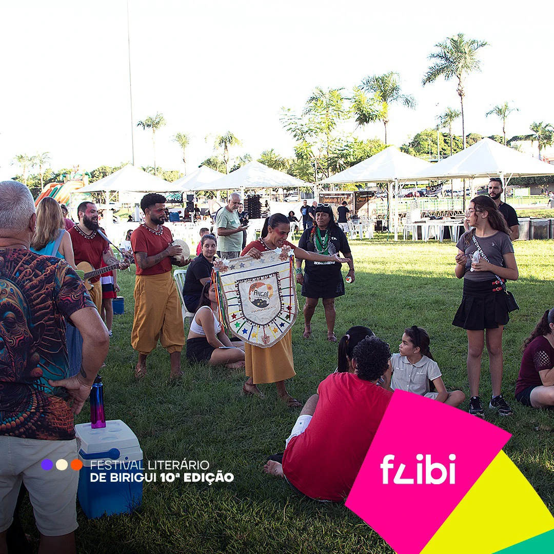 Roda de cultura popular no parque, ao ar livre: músicos com pandeiro e viola, uma mulher segurando um estandarte bordado e o público sentado na grama.