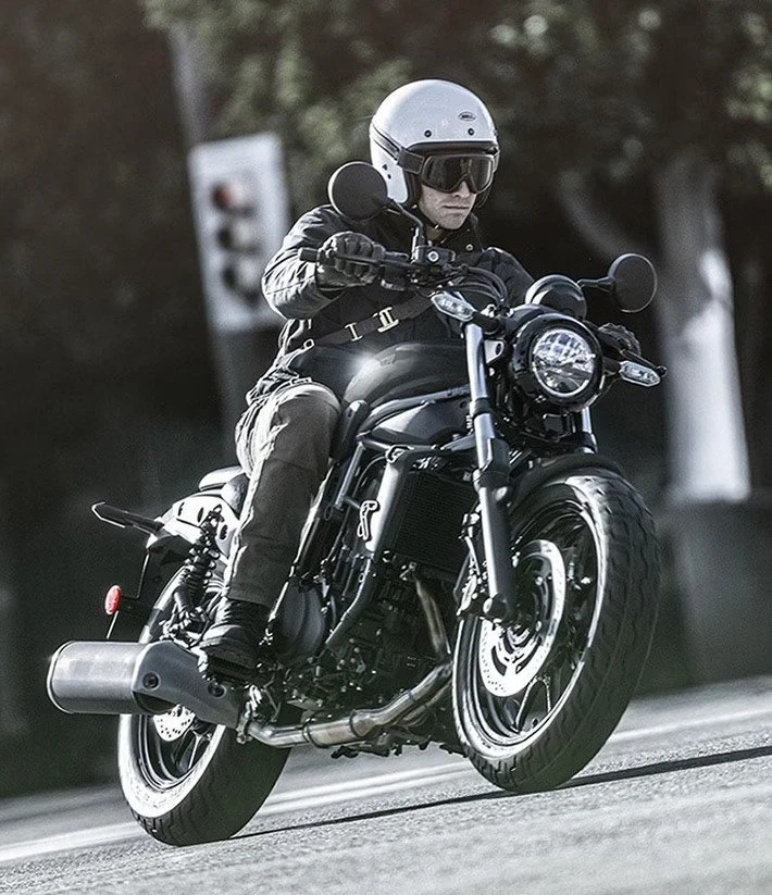 Piloto de capacete branco aberto e óculos de proteção conduzindo uma Kawasaki cruiser preta por uma via arborizada.