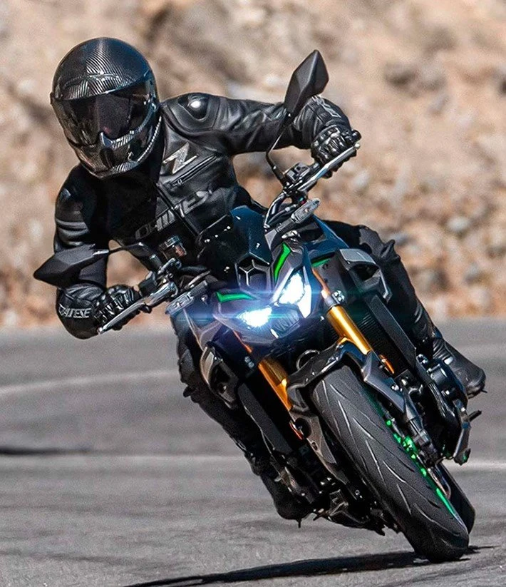 Piloto de macacão preto inclinado numa curva, de farol aceso, sobre uma Kawasaki naked de detalhes verdes.